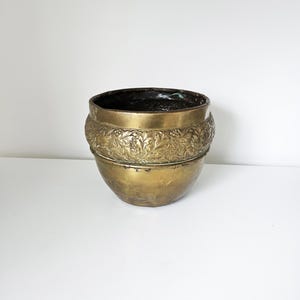 Op de afbeelding: Een vintage messing plantenbak met een donker interieur. De plantenbak heeft een afgeronde vorm met een decoratief bloemmotief rond het midden. Het messing heeft een warme, verouderde patina, wat bijdraagt aan de antieke charme.