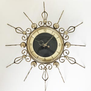 Op de afbeelding: Een vintage wandklok met een sterrenuitbarsting ontwerp. De klok heeft een goudkleurig metalen frame met decoratieve elementen en een zwarte wijzerplaat. De wijzers van de klok zijn zichtbaar en de klok is aan een witte muur bevestigd.