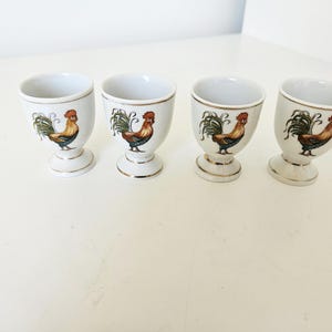 Op de afbeelding: Vier witte keramische eierdopjes met een gouden rand en een hanenontwerp. Elke beker heeft een gedetailleerde illustratie van een haan met rode, gele, groene en bruine kleuren. De eierdopjes staan op een wit oppervlak.