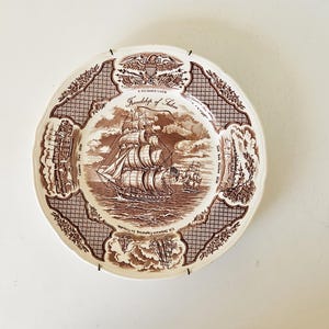Op de afbeelding: Een bruin en wit decoratief bord met een gedetailleerde illustratie van een zeilschip. Het bord heeft een decoratieve rand met de tekst "Friendship of Salem" en "E Pluribus Unum".