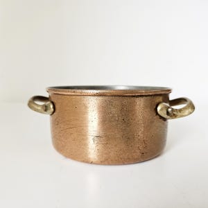 Op de afbeelding: Een kleine, koperkleurige kookpot met twee messing handvatten. De pot heeft een licht verouderd uiterlijk, met zichtbare slijtage op het oppervlak. De binnenkant van de pot lijkt een donkerder metaal te zijn. De pot staat op een witte ondergrond.