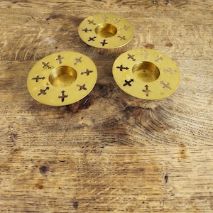 Op de afbeelding: Drie ronde koperen kandelaars met een centrale opening en een decoratieve rand van uitgesneden fleur-de-lis symbolen. Elk exemplaar heeft een gepolijste metalen afwerking en een platte, brede rand voor het plaatsen van een kaars.