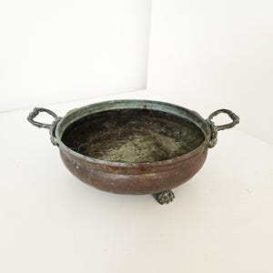 Pode incluir: Uma tigela de bronze antiga e desgastada, com duas alças ornamentadas e três pequenos pés. A tigela tem formato arredondado com interior escuro e exterior patinado, sugerindo idade e uso. A estética geral é rústica e vintage.