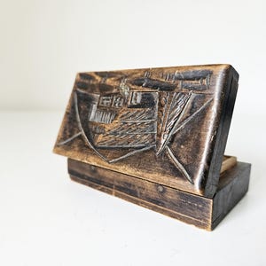 Op de afbeelding: Een kleine, open houten kist met een scharnierend deksel. Het deksel is ingewikkeld gesneden met een gedetailleerd ontwerp, mogelijk een gebouw of landschap. De kist is donkerbruin gekleurd en het interieur is zichtbaar.