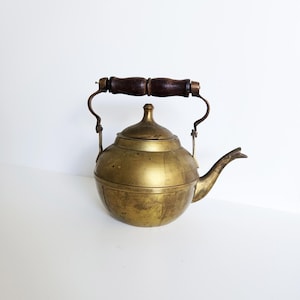 Op de afbeelding: Vintage messing theepot met een donkerhouten handvat. De theepot heeft een afgeronde body, een gebogen tuit en een deksel met een kleine knop. Het handvat is bevestigd met gebogen metalen steunen.