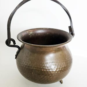 Op de afbeelding: Een gehamerde koperen ketel met een donkere, gebogen handgreep en drie kleine voetjes. De pot heeft een afgeronde body en een donker interieur. De handgreep is gemaakt van een donkerder metaal, wat contrasteert met het koper. Waarschijnlijk gebruikt om te koken of te decoreren.