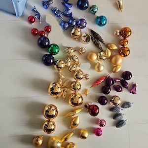 Op de afbeelding: Een verzameling diverse kerstballen in verschillende vormen, maten en kleuren, waaronder gouden, blauwe, rode, paarse en turquoise bollen, klokjes, druppels en dennenappelvormige decoraties verspreid over een wit oppervlak.