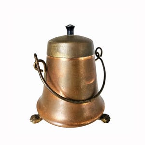 May include: A copper-colored metal pot with a matching lid featuring a black knob. The vessel has a curved metal handle attached to the sides and rests on three small feet shaped like animal paws, providing a stable base for the container.