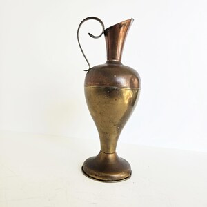 Op de afbeelding: Vintage messing kan met een gebogen handvat en een uitlopende tuit. De kan heeft een tweekleurige afwerking, met een donkerder bronzen bovenkant en een lichtere messing body. De basis is rond. Een decoratief stuk.