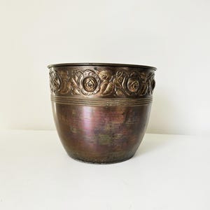 May include: A bronze-colored metal planter with a rounded shape. The top edge features a decorative band of embossed roses and foliage. The planter has a metallic sheen and sits on a white surface. A decorative item for indoor or outdoor use.