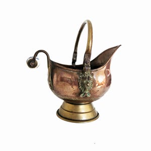 May include: A copper coal scuttle with a brass pedestal base and a curved handle. The vessel features a decorative brass lion head mount at the handle attachment point and a small, white, spherical knob on the side arm for tilting the container.