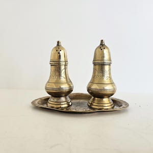 Op de afbeelding: Een vintage zout- en peperstrooier set van messing. De set bevat twee sierlijke strooiers met een decoratief ontwerp en een bijpassend ovaal dienblad. De strooiers hebben een klassieke, elegante vorm en zijn gemaakt van glanzend messing.