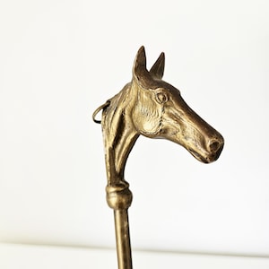Op de afbeelding: Een vintage messing paardenkop handvat met een gebogen steel. De paardenkop is gedetailleerd met een getextureerde afwerking en een kleine ring aan de bovenkant. De steel heeft een decoratief bolvormig gedeelte.