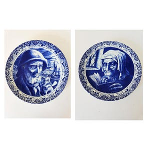 Op de afbeelding: Twee blauw-witte decoratieve borden. Eén bord toont een man met baard en hoed die een pijp rookt, met zeilboten op de achtergrond. Het andere bord toont een oudere vrouw die een boek leest. Beide borden hebben sierlijke, patroonranden.