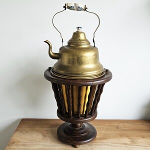 Op de afbeelding: Een vintage messing theepot met een decoratieve handgreep en een houten voet. De theepot heeft een gebogen tuit en een deksel met een kleine, decoratieve knop. De houten voet heeft een donkere afwerking en een uniek, open ontwerp. De theepot is goudkleurig.