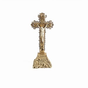 Puede incluir: Un crucifijo de pie con una cruz de metal en tono dorado antiguo, adornada con intrincados filigranas y una figura blanca central de Jesús. La cruz está montada sobre una base cuadrada ancha decorada con patrones florales y de hojas en relieve.