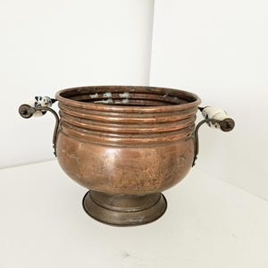Op de afbeelding: Een vintage koperen pot met een ronde body en een voetstuk. De pot heeft twee handvatten met witte keramische uiteinden versierd met blauwe bloempatronen. Het koper heeft een warme, verouderde patina.