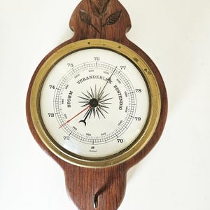 Könnte beinhalten: Ein Vintage-Barometer aus Holz mit einem messingfarbenen Ring und einem weißen Zifferblatt. Das Zifferblatt zeigt Zahlen, eine Nadel und die Wörter "STORM" und "BESTENDIG". Der Holzrahmen hat eine dekorative Ober- und Unterseite mit einem Haken.