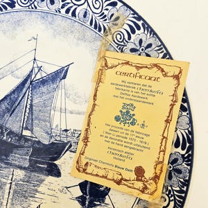 Op de afbeelding: Blauw en witte Delfts aardewerk bord met een zeilboot scène en bloemenrand. Een certificaat bevestigt de authenticiteit en herkomst van Chemkefa Blauw Delft, Holland.
