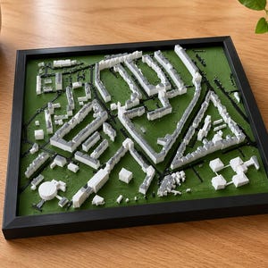 May include: A three-dimensional square wall art piece featuring a white architectural model of a city neighborhood set against a textured green background, all enclosed within a thin black frame. The model displays various building shapes.