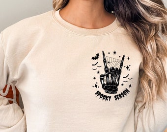 May include: A beige sweatshirt featuring a black graphic of a skeleton hand making a rock and roll sign. The graphic also includes a spider web, bats, and the text "Spooky Season".