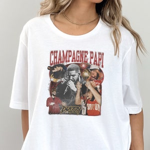 May include: White t-shirt with a graphic print of a group of people, including a man with a microphone, and the text "Champagne Papi" and "Drizzy".
