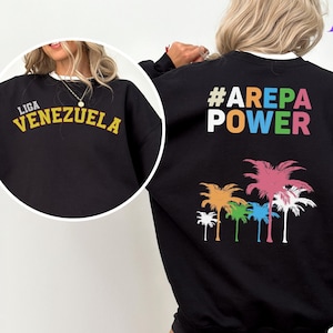 May include: Black sweatshirt with a colorful palm tree graphic and the text "#AREPA POWER" on the back. The front of the sweatshirt has the text "LIGA VENEZUELA" in yellow.