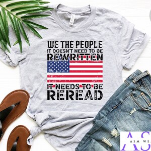 以下が含まれることがあります： 「WE THE PEOPLE IT DOESN'T NEED TO BE REWRITTEN IT NEEDS TO BE REREAD」というテキストと、くたびれたアメリカ国旗のデザインが施されたグレーのTシャツ。