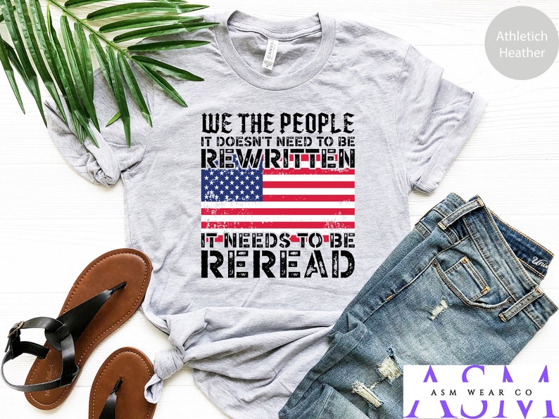 以下が含まれることがあります： 「WE THE PEOPLE IT DOESN'T NEED TO BE REWRITTEN IT NEEDS TO BE REREAD」というテキストと、くたびれたアメリカ国旗のデザインが施されたグレーのTシャツ。