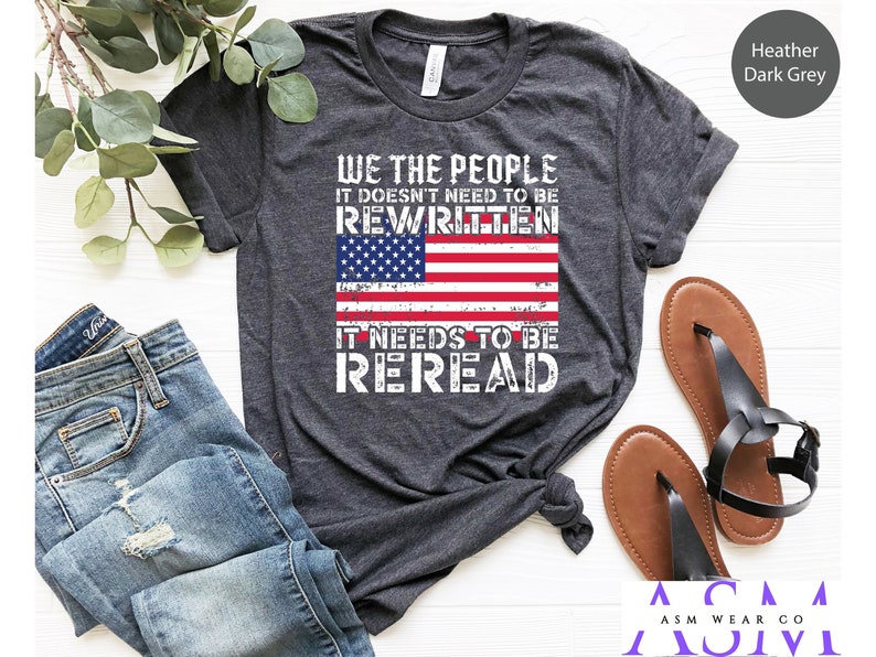 以下が含まれることがあります： ヘザーダークグレーのTシャツに、ダメージ加工されたアメリカ国旗のグラフィックと「WE THE PEOPLE IT DOESN'T NEED TO BE REWRITTEN IT NEEDS TO BE REREAD」のテキストがプリントされています。
