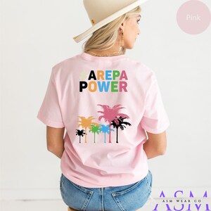 May include: Pink t-shirt with the text "AREPA POWER" in colorful letters and a graphic of palm trees on the back.