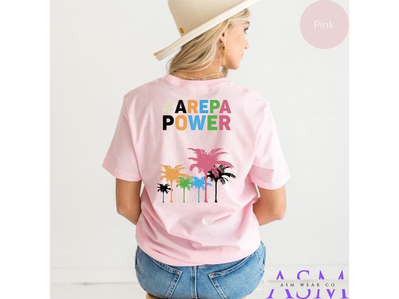 May include: Pink t-shirt with the text "AREPA POWER" in colorful letters and a graphic of palm trees on the back.