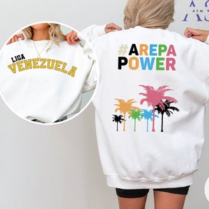 May include: White sweatshirt with the text "LIGA VENEZUELA" on the front and "#AREPA POWER" with a row of colorful palm trees on the back.