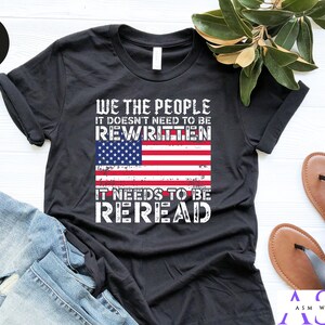 以下が含まれることがあります： 「We the People It Doesn't Need to Be Rewritten It Needs to Be Reread」というテキストと、くたびれたアメリカ国旗のグラフィックがプリントされた黒いTシャツ。