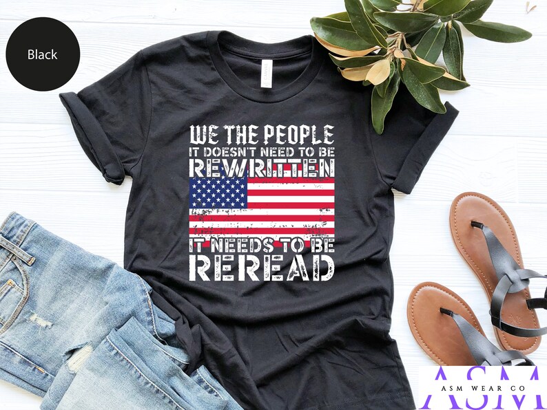 以下が含まれることがあります： 「We the People It Doesn't Need to Be Rewritten It Needs to Be Reread」というテキストと、くたびれたアメリカ国旗のグラフィックがプリントされた黒いTシャツ。