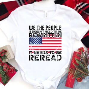 以下が含まれることがあります： 「We the People It doesn't need to be rewritten It needs to be reread」というテキストと、くたびれたアメリカ国旗のグラフィックがプリントされた白いTシャツ。