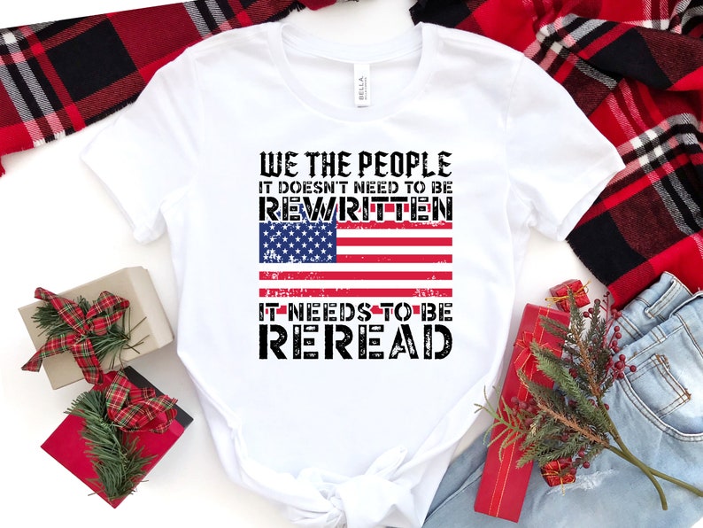 以下が含まれることがあります： 「We the People It doesn't need to be rewritten It needs to be reread」というテキストと、くたびれたアメリカ国旗のグラフィックがプリントされた白いTシャツ。