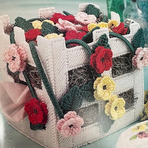 Puede incluir: Una cesta blanca tejida adornada con flores rojas, rosas y amarillas de ganchillo y hojas verdes. La cesta está llena de una mezcla de las mismas flores y vegetación, creando un arreglo floral. Un lazo rosa está unido al costado.