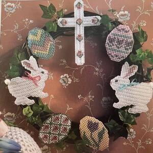 May include: A handmade Easter wreath featuring a white cross, decorative eggs, and two white bunnies with bows. The wreath is adorned with green leaves and set against a floral background, perfect for Easter decor.