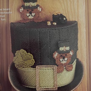 May include: A handmade St. Patrick's Day themed top hat in green and gold. The hat is decorated with a shamrock, a decorative buckle, and two teddy bears wearing miniature top hats. A small pot of gold and gold coins are also present.