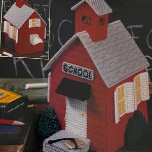 Op de afbeelding: Een rode schoolgebouw-vormige tissueboxhoes met een grijs dak en het woord "SCHOOL" boven de deur. De ramen hebben witte luiken en gele accenten. Een klein grijs boek en een grijze bank staan voor het schoolgebouw.