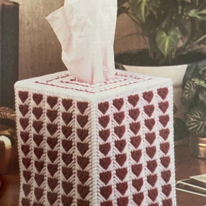 Puede incluir: Una funda de caja de pañuelos cuadrada con fondo blanco y un patrón de corazones burdeos. La parte superior de la caja tiene un borde rojo, y los pañuelos blancos sobresalen del centro. La caja está sobre una superficie de madera.