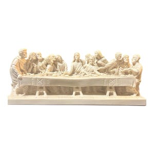 May include: A cream-colored sculptural figurine depicting The Last Supper, featuring thirteen figures seated at a long rectangular table with a draped cloth, all rendered in a monochromatic, detailed relief style on a solid base.
