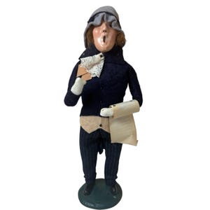 May include: A vintage Christmas carol singer figurine. The figure wears a dark blue coat, striped pants, and a gray hat. It holds a sheet of paper and a card with a white lace collar. The figurine stands on a green base.