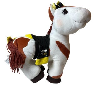 May include: A soft toy horse with a white body, brown markings, and a brown mane and tail. The horse has a black saddle with yellow trim and a bee emblem. It has a smiling face and is wearing a yellow bow.