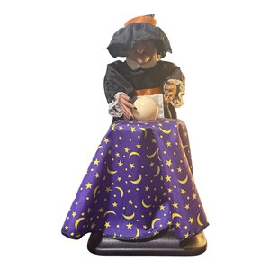 May include: A witch figurine wearing a black hat with an orange band, a black top with white lace cuffs, and a purple skirt featuring a yellow moon and star pattern. The figure has hands positioned over a smooth, round, off-white crystal ball.