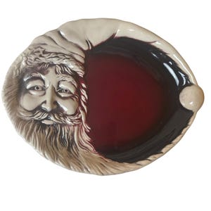 May include: An oval ceramic dish featuring a sculpted Santa Claus face on the left side with a detailed beard and hat. The right side of the dish is a smooth, deep red glazed surface, and the rim is finished with a cream-colored border.