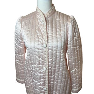 Puede incluir: Una bata acolchada de color rosa claro con cuello alto y lunares blancos. La bata tiene una parte delantera abotonada con botones blancos y un ribete blanco decorativo a lo largo de los bordes. Las mangas son abullonadas en los hombros.