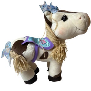Può includere: Un cavallo di peluche con corpo bianco, macchie marroni e criniera marrone. Ha una sella blu e viola con un ciondolo a farfalla e nastri blu sulla coda e sulla testa. Il cavallo ha un volto sorridente ed è progettato per bambini.
