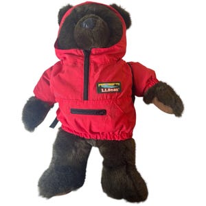 Puede incluir: Un oso de peluche marrón con una chaqueta roja con capucha, cremallera negra y el logotipo "L.L.Bean". El oso tiene pelaje marrón oscuro y está de pie. La chaqueta tiene un cordón en la parte inferior.